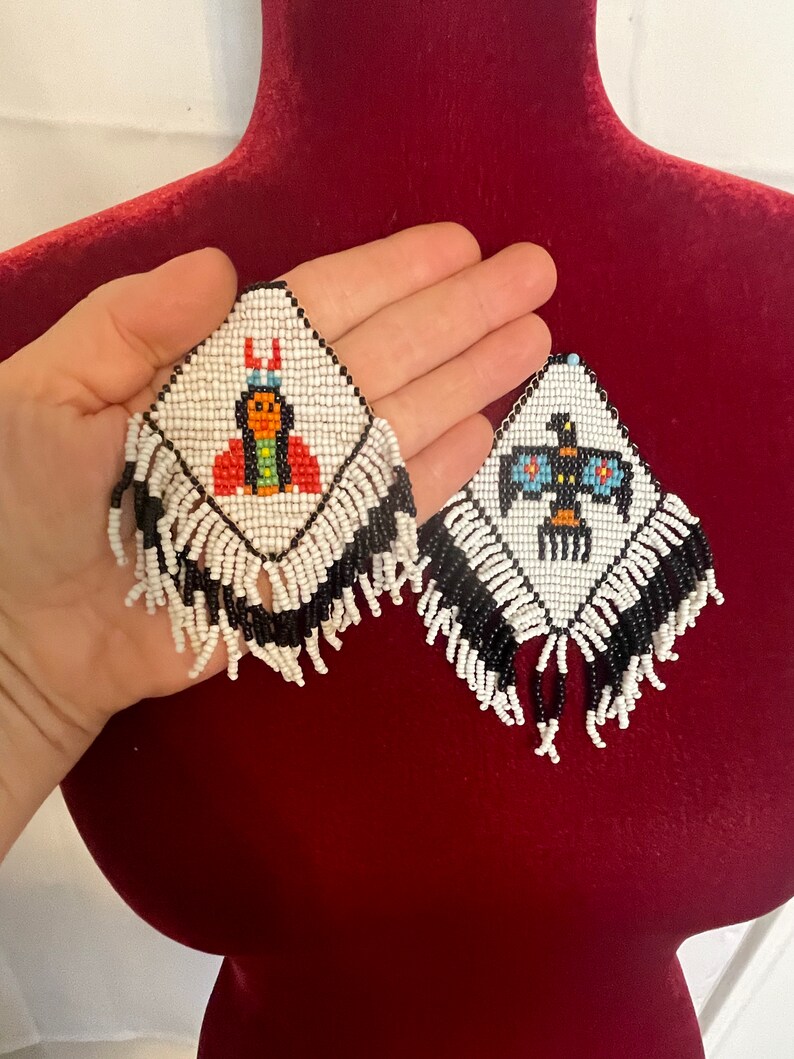 Puede incluir: Un par de pendientes de cuentas blancas y negras con flecos. Un pendiente presenta una imagen de cuentas de una mujer nativa americana con ropa tradicional, mientras que el otro pendiente presenta una imagen de cuentas de un p&aacute;jaro con las alas extendidas.