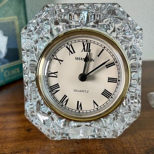 May include: A clear, faceted crystal quartz clock with a gold-tone bezel and a cream-colored face. The clock face features black Roman numerals and the brand name "SHANNON". The clock is set on a wooden surface.