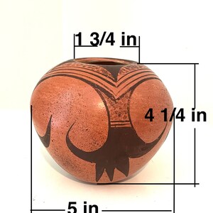 以下が含まれることがあります： 黒い幾何学模様が施された、小さな丸いテラコッタ製の壺。壺の高さは10.8cm、幅は12.7cm、開口部の直径は4.4cmです。
