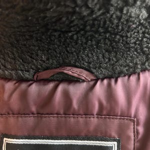 Puede incluir: Primer plano del interior de una chaqueta burdeos con cuello de piel sint&eacute;tica negra. La chaqueta tiene una etiqueta con el texto "TRADE MARK 1973 VINTAGE ESTABLISHED". Se ve un peque&ntilde;o lazo cerca del cuello.