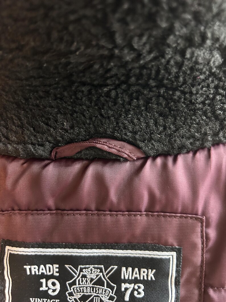 Puede incluir: Primer plano del interior de una chaqueta burdeos con cuello de piel sint&eacute;tica negra. La chaqueta tiene una etiqueta con el texto "TRADE MARK 1973 VINTAGE ESTABLISHED". Se ve un peque&ntilde;o lazo cerca del cuello.