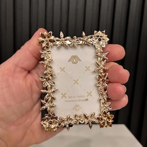 May include: Gold-toned picture frame with a floral design and clear rhinestones. The frame is 2x3 inches or 5x8 centimetres. The text "Presented by Frame" is printed on the frame.