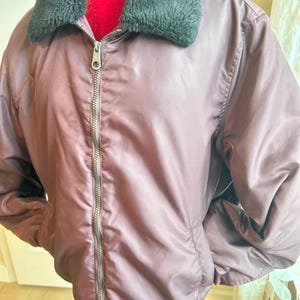 Puede incluir: Una chaqueta bomber malva con cuello de piel sint&eacute;tica negra y cremallera plateada. La chaqueta tiene dos bolsillos delanteros y un dobladillo acanalado. La chaqueta est&aacute; hecha de un material liso y brillante.