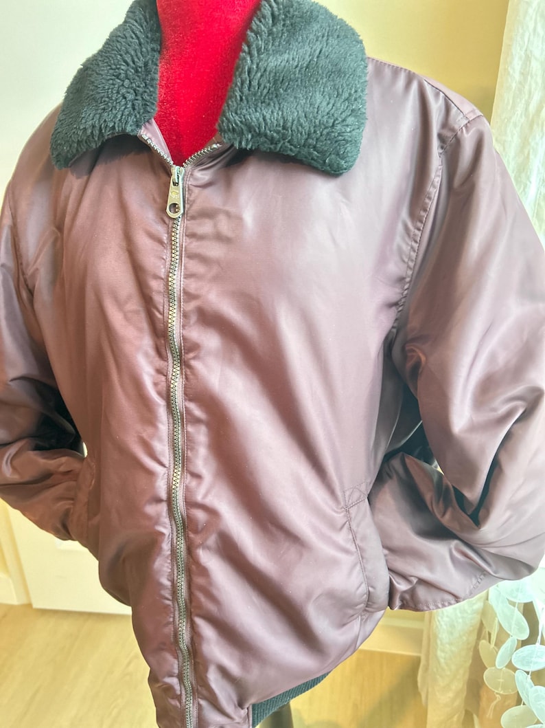Puede incluir: Una chaqueta bomber malva con cuello de piel sint&eacute;tica negra y cremallera plateada. La chaqueta tiene dos bolsillos delanteros y un dobladillo acanalado. La chaqueta est&aacute; hecha de un material liso y brillante.