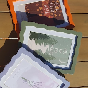 May include: Three decorative picture frames in orange, green, and lavender. Each frame has a wavy edge and holds a different print. The prints feature fall leaves, a forest scene, and lavender sprigs. The frames are stacked on a wooden surface.