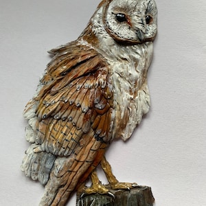 Könnte beinhalten: Handbemalte Eulen-Skulptur mit detaillierter, strukturierter Oberfläche. Die Eule hat ein weißes Gesicht, braune und beige Flügel und sitzt auf einem kleinen Holzstumpf. Dekorativer Artikel für die Inneneinrichtung mit rustikaler Ästhetik.