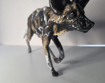 Puede incluir: Figura detallada de un licaón, también conocido como perro salvaje africano, en posición de caminar. La escultura presenta una representación realista con una mezcla de marcas marrones, negras y blancas. El perro tiene orejas grandes y redondeadas y una expresión concentrada.