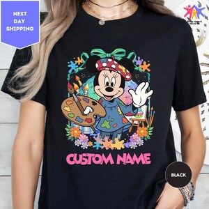 A black t-shirt with a colorful graphic of Minnie Mouse holding a paintbrush and surrounded by flowers and splatters. The text "CUSTOM NAME" is displayed below the graphic.