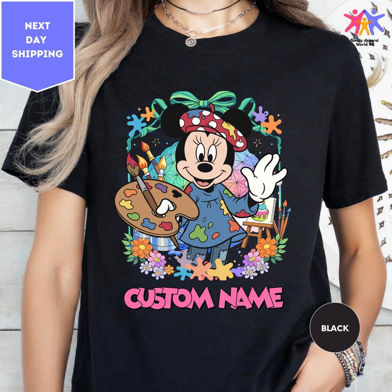 A black t-shirt with a colorful graphic of Minnie Mouse holding a paintbrush and surrounded by flowers and splatters. The text "CUSTOM NAME" is displayed below the graphic.