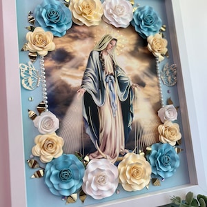 May include: A framed religious artwork featuring a central image of a figure in flowing robes, surrounded by paper flowers in shades of blue, cream, and white. Decorative butterflies and pearl accents add detail to the light blue background.