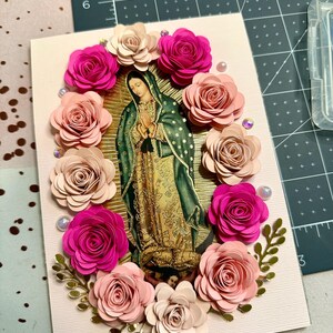 May include: A handmade greeting card featuring an image of Our Lady of Guadalupe, surrounded by paper roses in shades of pink and cream. The card is embellished with pearls and gold accents, creating a devotional piece.