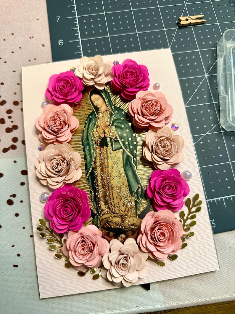 May include: A handmade greeting card featuring an image of Our Lady of Guadalupe, surrounded by paper roses in shades of pink and cream. The card is embellished with pearls and gold accents, creating a devotional piece.
