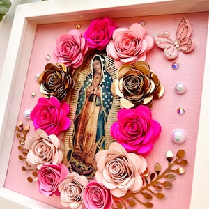 May include: A framed artwork featuring a religious image surrounded by paper roses in shades of pink and gold. A butterfly and decorative accents complete the design. The artwork is set against a pink background within a white frame.