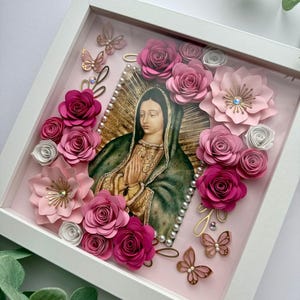 May include: A framed image of Our Lady of Guadalupe surrounded by pink and white paper flowers and gold butterflies. The frame is white and the background is pink.