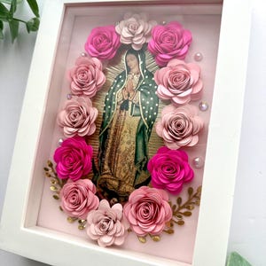 May include: A framed religious image of Our Lady of Guadalupe, surrounded by pink paper roses and gold accents. The image is set against a light pink background within a white frame, creating a layered, dimensional effect. Decorative beads add a touch of sparkle.