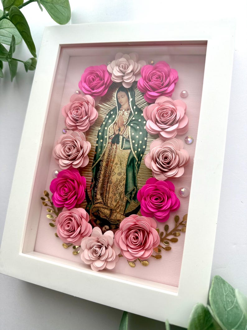 May include: A framed religious image of Our Lady of Guadalupe, surrounded by pink paper roses and gold accents. The image is set against a light pink background within a white frame, creating a layered, dimensional effect. Decorative beads add a touch of sparkle.