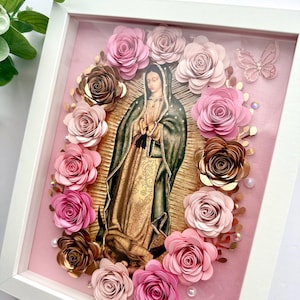 May include: A framed image of the Virgin Mary surrounded by pink and gold paper flowers. The image is set against a pink background and is framed in white.
