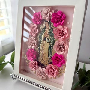 May include: A framed artwork featuring an image of Our Lady of Guadalupe, surrounded by paper roses in shades of pink. The artwork is encased in a white frame and displayed on a silver stand. The image has a gold halo and is set against a pink background.