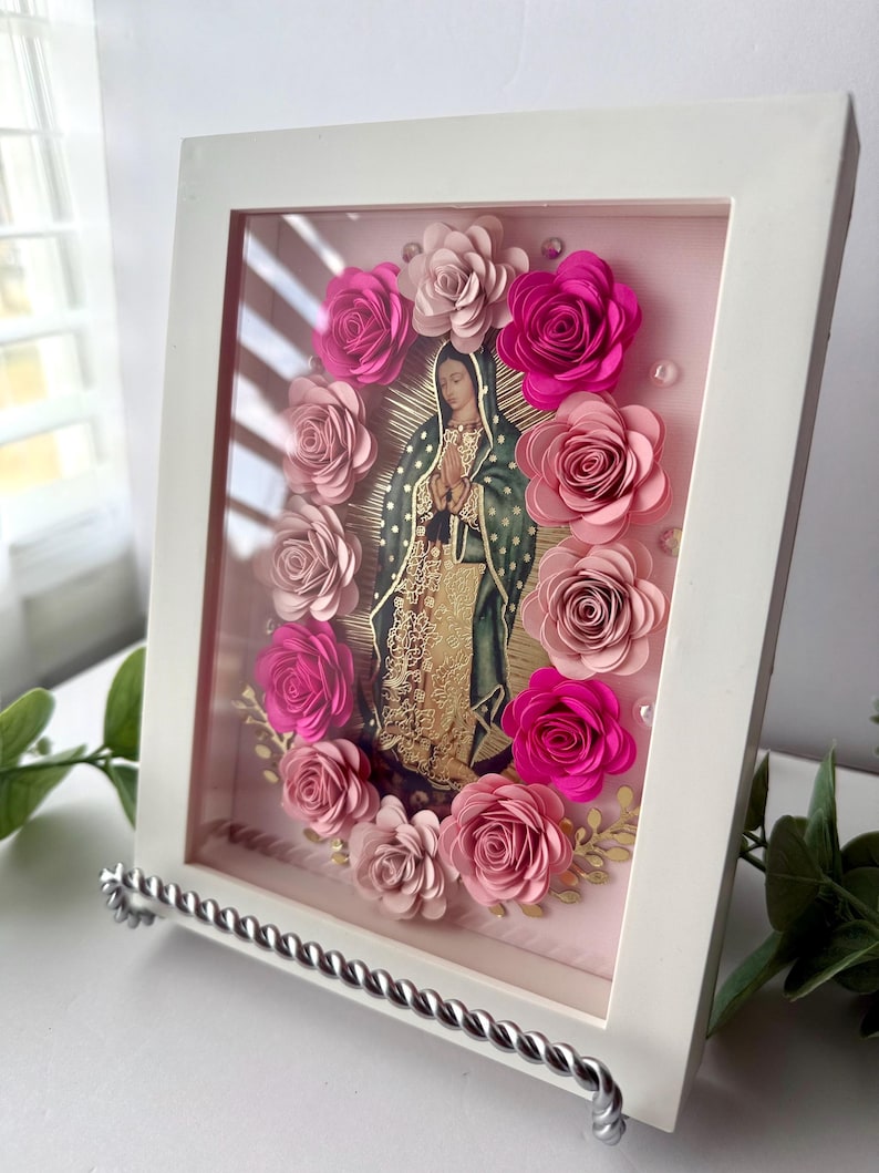 May include: A framed artwork featuring an image of Our Lady of Guadalupe, surrounded by paper roses in shades of pink. The artwork is encased in a white frame and displayed on a silver stand. The image has a gold halo and is set against a pink background.