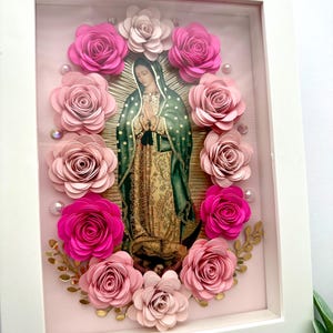 May include: A framed religious artwork featuring an image of the Virgin Mary surrounded by pink paper roses. The artwork is set against a light pink background with gold accents and a white frame.