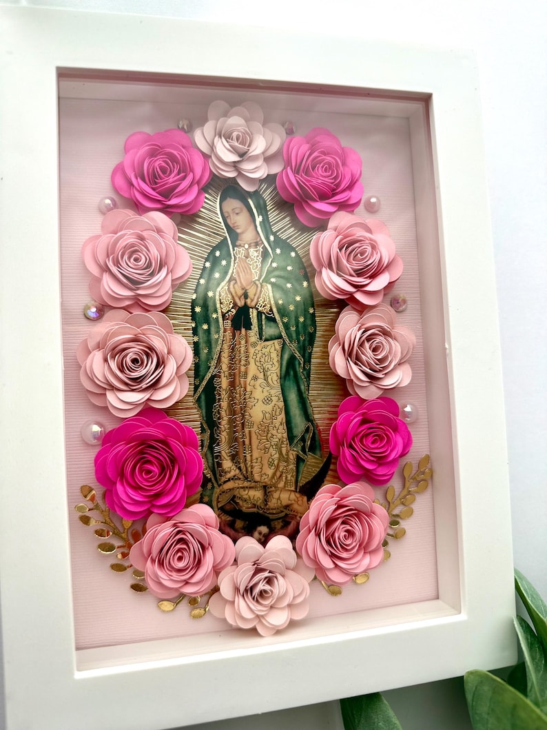 May include: A framed religious artwork featuring an image of the Virgin Mary surrounded by pink paper roses. The artwork is set against a light pink background with gold accents and a white frame.