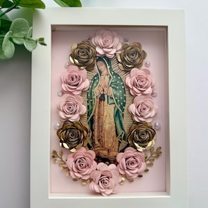 May include: A framed image of Our Lady of Guadalupe surrounded by pink and gold paper flowers. The image is on a pink background.