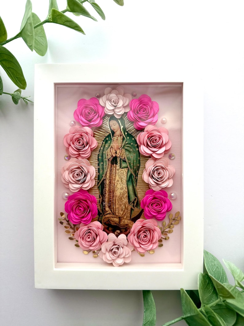 May include: A framed religious artwork featuring an image of the Virgin Mary surrounded by paper roses in shades of pink and fuchsia. The artwork is set against a pink background and framed in a white rectangular frame.