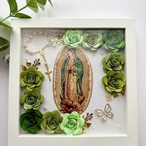 May include: A white frame with a picture of the Virgin Mary inside. The frame is decorated with green paper flowers, a gold butterfly, and a rosary.