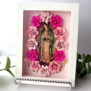 May include: A framed religious image of Our Lady of Guadalupe, surrounded by pink paper roses. The artwork features a white frame and a silver stand. The image of the Virgin Mary is in the center, with a pink background.