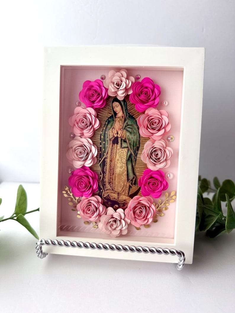 May include: A framed religious image of Our Lady of Guadalupe, surrounded by pink paper roses. The artwork features a white frame and a silver stand. The image of the Virgin Mary is in the center, with a pink background.