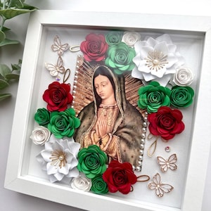 May include: A white framed picture with a religious image of the Virgin Mary surrounded by paper flowers in red, green, and white. The flowers are layered and have a 3D effect.