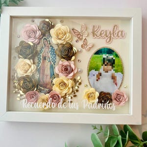 May include: A framed shadow box with a religious theme. It features a central image of a religious figure, surrounded by paper flowers in shades of pink, gold, and cream. The name "Keyla" is displayed, along with the text "Recuerdo de tus Padrinos".
