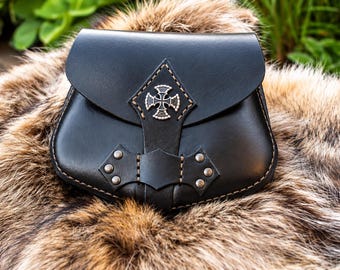 Puede incluir: Un bolso de cuero negro con cierre de solapa y un emblema de cruz plateado centrado en la parte delantera. El bolso presenta costuras visibles a lo largo de los bordes y cuatro pequeños remaches metálicos plateados en el panel frontal inferior.