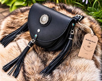 Puede incluir: Un bolso de cuero negro con un adorno circular plateado tipo concho, detalles de piedra turquesa y largas borlas de cuero negro. Una etiqueta de papel marrón atada con cordel muestra el texto Nathaly Fox & Co.