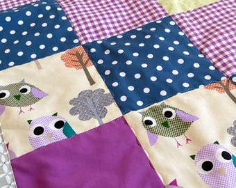 Peut inclure: Une courtepointe patchwork avec des carrés de différents motifs et couleurs. La courtepointe comprend des carrés avec des motifs de hiboux et d'arbres, des pois, du vichy et du violet uni. La bordure est vert clair avec un motif floral blanc.