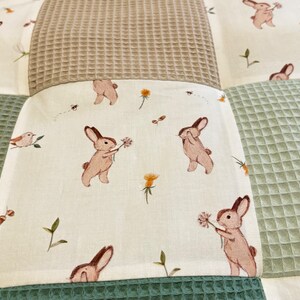 Puede incluir: Una manta de beb&eacute; de patchwork con un patr&oacute;n de tejido de gofre verde, marr&oacute;n y blanco. La tela blanca presenta un patr&oacute;n repetido de conejos marrones que sostienen flores.