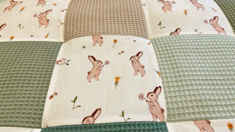 Puede incluir: Una manta de beb&eacute; de patchwork con un patr&oacute;n de tejido de gofre verde, marr&oacute;n y blanco. La tela blanca presenta un patr&oacute;n repetido de conejos marrones que sostienen flores.