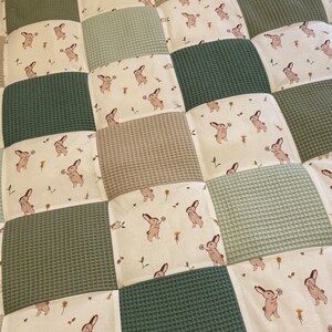 Puede incluir: Una alfombra de juego para beb&eacute;s de patchwork con fondo crema y cuadrados de textura de gofre verde. Los cuadrados crema presentan un patr&oacute;n repetitivo de conejos marrones que sostienen flores.