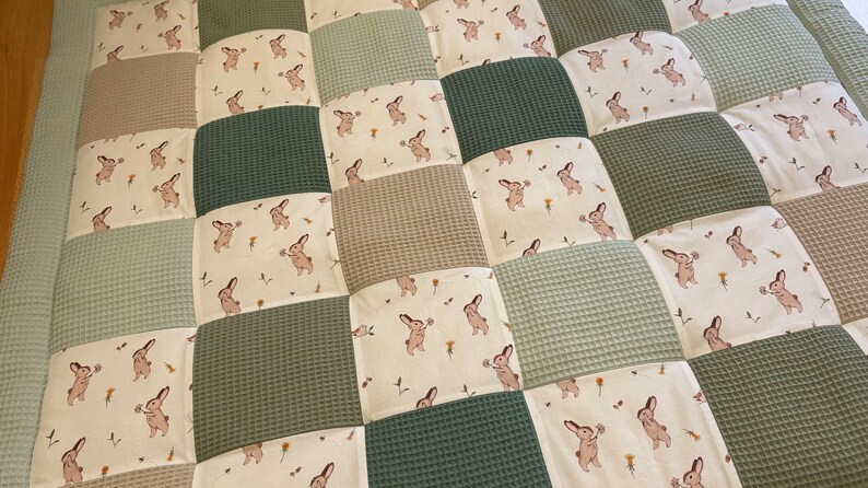 Puede incluir: Una alfombra de juego para beb&eacute;s de patchwork con fondo crema y cuadrados de textura de gofre verde. Los cuadrados crema presentan un patr&oacute;n repetitivo de conejos marrones que sostienen flores.