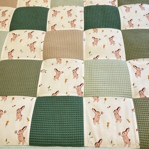 Puede incluir: Una alfombra de juego para beb&eacute;s de patchwork con un esquema de color verde, beige y blanco. La alfombra de juego est&aacute; hecha de tela suave acolchada y presenta un patr&oacute;n repetitivo de conejos con flores.