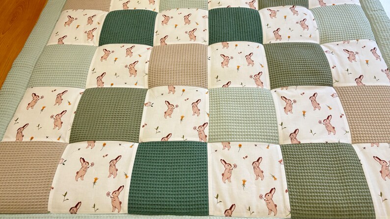 Puede incluir: Una alfombra de juego para beb&eacute;s de patchwork con un esquema de color verde, beige y blanco. La alfombra de juego est&aacute; hecha de tela suave acolchada y presenta un patr&oacute;n repetitivo de conejos con flores.