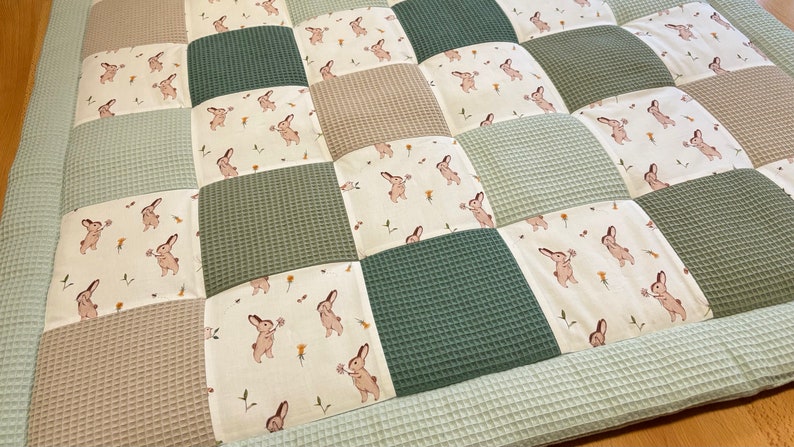 Puede incluir: Una alfombra de juego de patchwork en color verde, beige y blanco. La alfombra est&aacute; hecha de tela acolchada con textura de gofre. La tela presenta un patr&oacute;n repetido de conejos que sostienen flores.