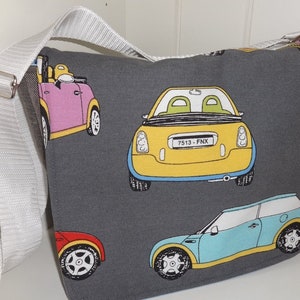 Könnte beinhalten: Eine graue Canvas-Messenger-Tasche mit einem weißen Trageriemen. Die Tasche hat ein sich wiederholendes Muster aus bunten Mini-Cooper-Autos im Cartoon-Stil. Ein Auto hat das Kennzeichen "7513-FNX".
