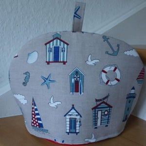 Könnte beinhalten: Ein beiger Teewärmer mit maritimem Design. Er zeigt Illustrationen von Strandhütten, Segelbooten, Ankern und Seesternen in Blau und Rot. Weiße Möwen und Wolken sind ebenfalls auf dem Stoff abgebildet.