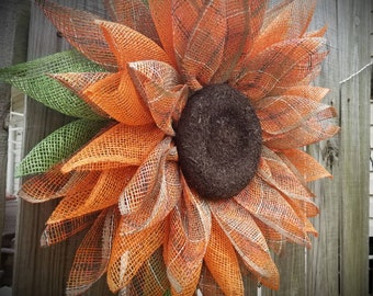 May include: A large orange and green mesh sunflower wreath with a brown center. The wreath is made of burlap and mesh and is perfect for fall decor.