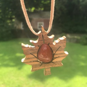 May include: A wooden maple leaf pendant with a red carnelian stone in the center. The pendant is on a brown leather cord.
