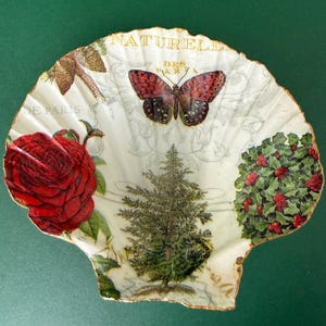 May include: A decorative seashell with a painted design. The design includes a red rose, a butterfly, a small evergreen tree, and a holly bush with red berries. The shell has gold trim and the words "NATUREL" and "DE PARIS" are visible.