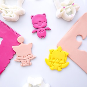 Op de afbeelding: Een verzameling witte plastic koekjesvormpjes en bijbehorende figuren van roze, perzikkleurige en gele klei. De vormen tonen gestileerde wezens met vleugels en puntige oren, naast platte stukken gekleurde klei en een witte uitsteekvorm.