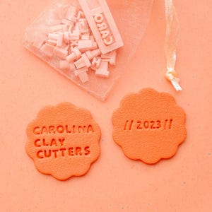 Puede incluir: Una colección de cortadores de arcilla y accesorios de color naranja. La imagen presenta una pequeña bolsa de herramientas de arcilla, dos sellos cortadores de arcilla naranja con el texto "CAROLINA CLAY CUTTERS" y "// 2023 //".