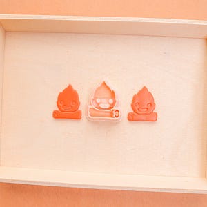 May include: Three orange-colored, campfire-shaped cookie cutters sit inside a light-colored wooden tray. The cookie cutters have a cartoon-like design, with a smiling face and flames. The tray has cut-out handles.